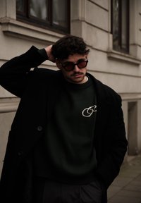 Manteau noir sur un pull vert avec un motif brodé, associé à des lunettes de soleil foncées. Arrière-plan urbain avec des couleurs et textures atténuées.