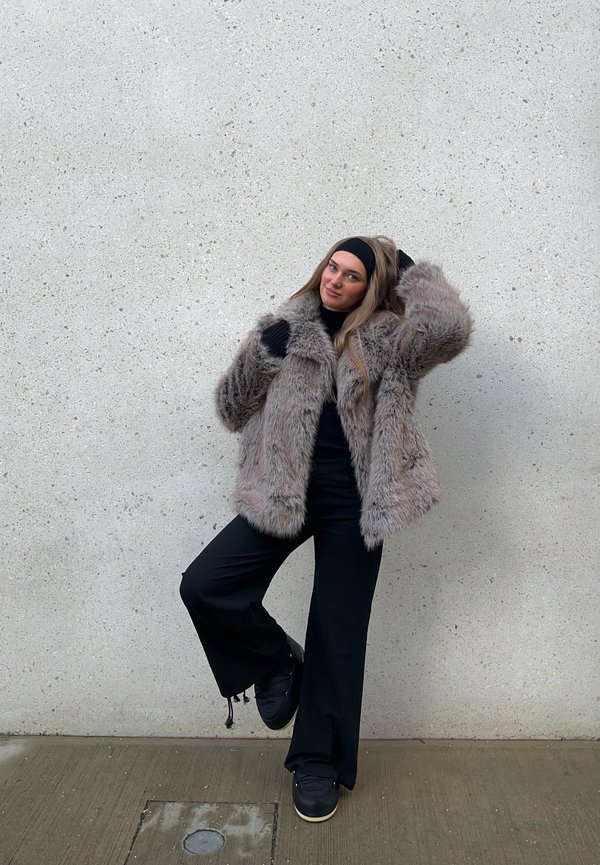 Umělé kožich ve světle hnědé barvě s hustou texturou, nošený přes černý top a rozšířené černé kalhoty. Černé rukavice a boty dokončují vzhled.
