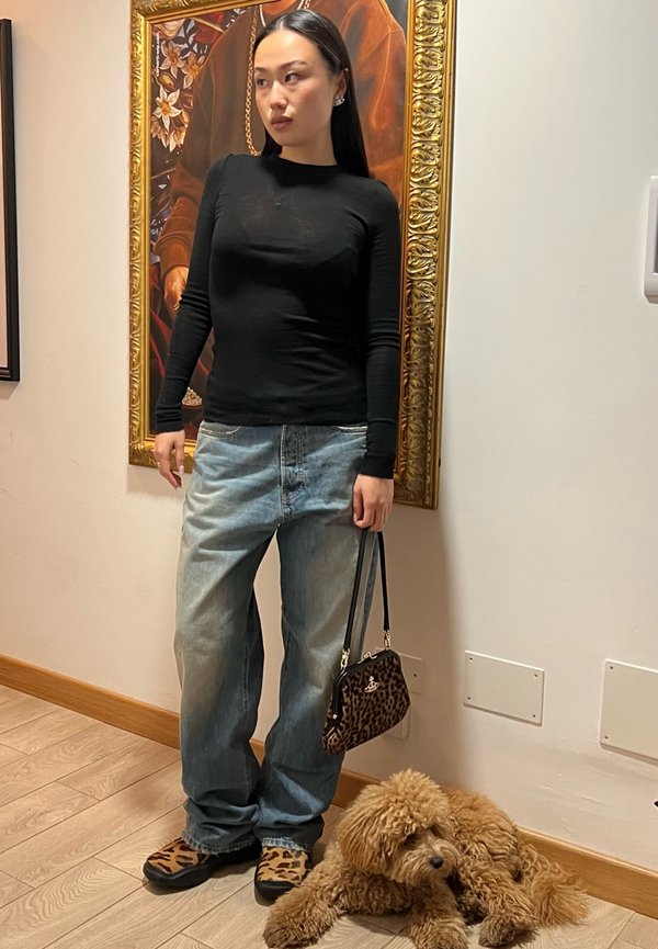 Sort langærmet top, lyseblå løse jeans, leopardmønstrede sko og håndtaske. En brun hund ligger på gulvet ved siden af personen.