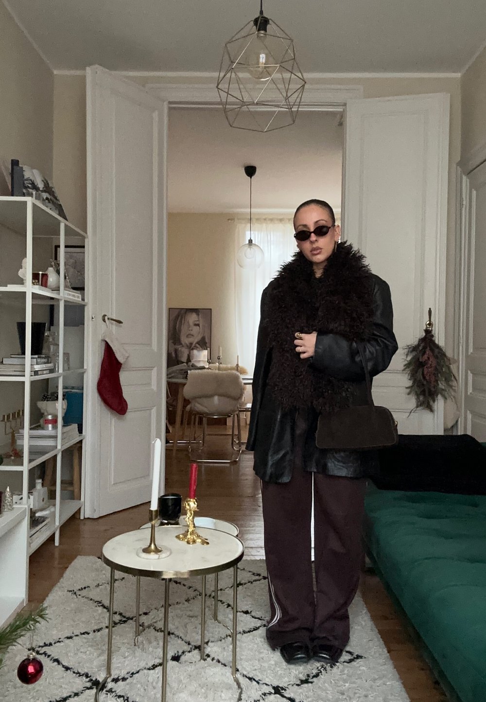 Personne portant une grande veste en cuir sombre à col en fourrure, des lunettes de soleil et un pantalon marron, debout dans un salon décoré avec une petite table ronde.