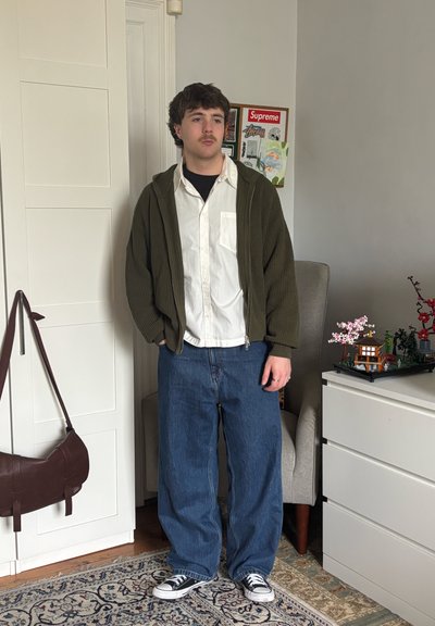 Joven con bigote que lleva un suéter de cremallera oliva, camisa blanca, vaqueros anchos azules y zapatillas negras, de pie en una habitación con una alfombra y un armario.