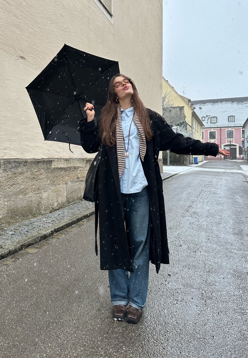 Young woman wearing glasses and a long coat holds a black umbrella while standing on a wet street with light snowfall.