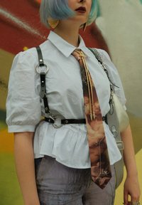 Hvid bluse med pufærmer og peplum-design, sort læderharness med metalringe, og et mønstret slipse med et klassisk print. Sølvtilbehør.