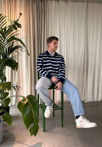 Sudadera de forro polar a rayas en navy y blanco, jeans de lavado claro, zapatillas blancas, taburete verde, fondo con cortinas suaves y plantas en maceta.