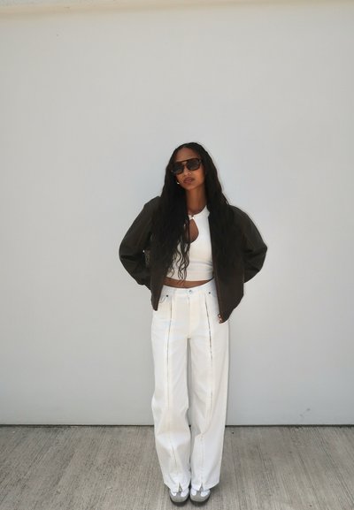 Chaqueta bomber marrón, top corto blanco y pantalones anchos blancos con costuras en contraste. Gafas de sol oscuras y cabello texturizado y ondulado.