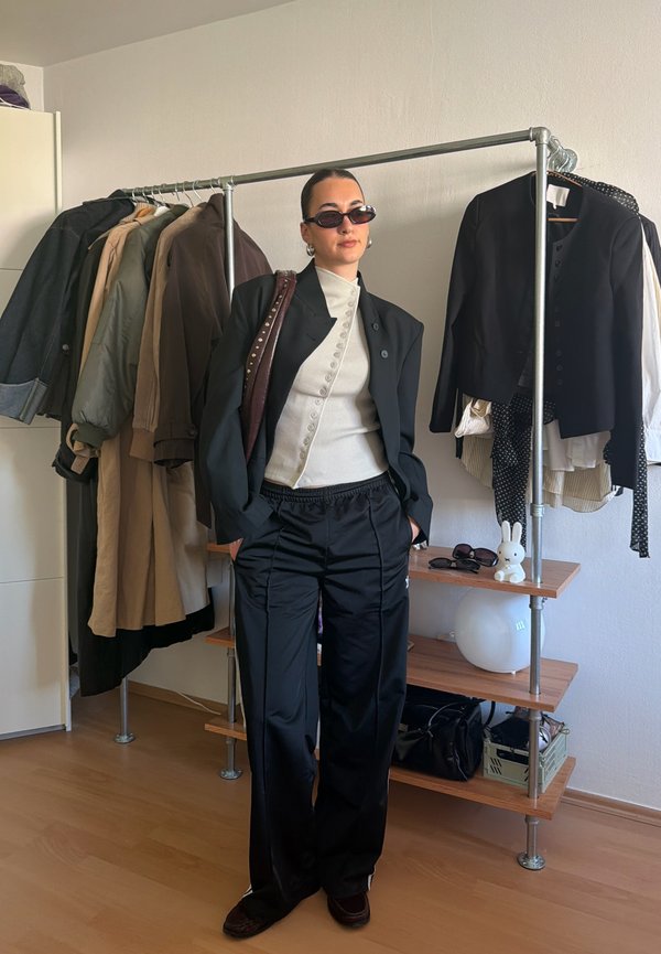 Persona que lleva gafas de sol negras, blazer negro, top blanco abotonado y pantalones negros, de pie junto a un perchero con chaquetas y estantes.