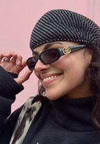 Jeune femme souriante ajustant des lunettes de soleil Gucci noires, portant un bonnet tricoté rayé, un col roulé noir et un accessoire à motif de fourrure.