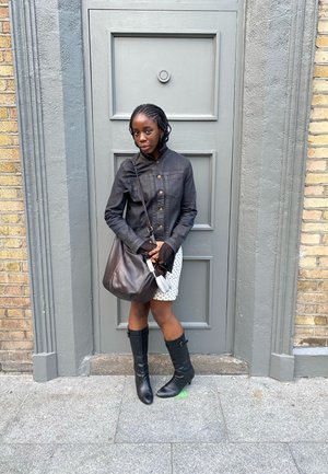 Joven con el cabello trenzado, lleva una chaqueta negra, falda blanca con lunares, botas negras y sostiene una gran bolsa marrón junto a una puerta gris.