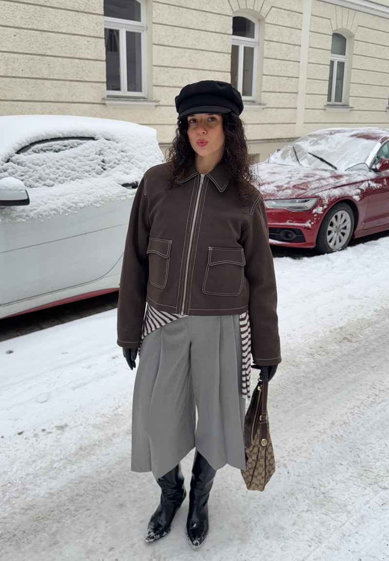 Veste marron à fermeture éclair avec coutures blanches, culottes plissées grises, cheveux noirs bouclés, bottes noires et sac à main à motifs, debout sur la neige.