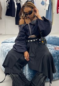 Zwarte cropped jasje met off-shoulder ontwerp, brede zwarte broek, gelaagde texturen en een zonnebril. Zittend op een blauwe textuurpouf.