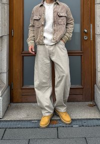 Personne debout portant une veste en velours côtelé beige, un pull blanc, un pantalon ample beige et des bottes jaunes devant une porte en bois avec des panneaux en verre dépoli.