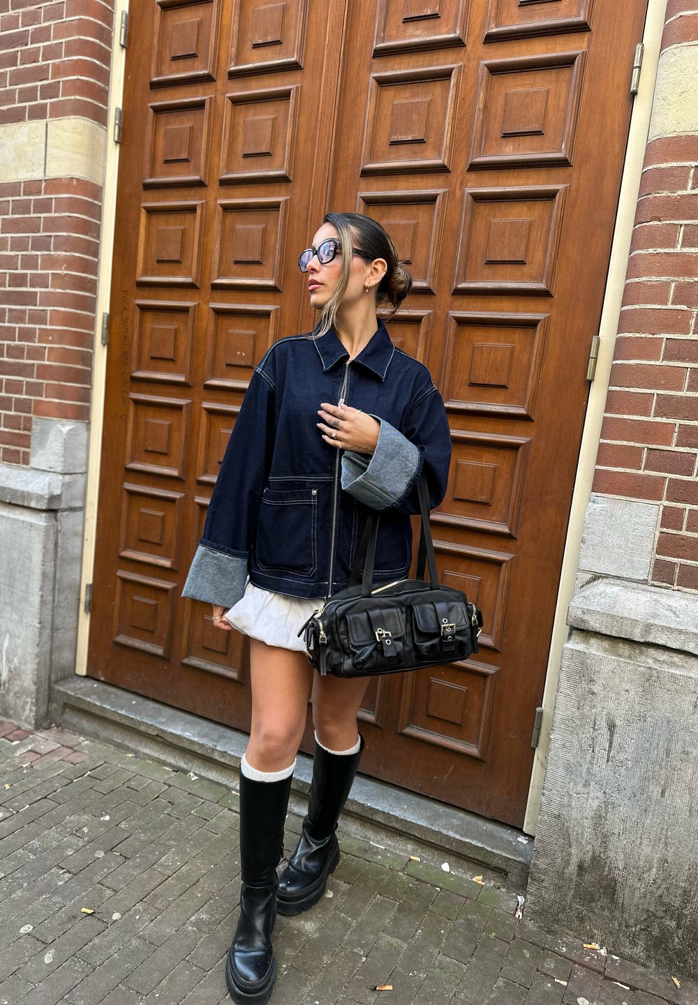 Donkerdenimjack met grijze manchetten, gedragen over een witte rok. Zwarte handtas met meerdere zakken. Zwarte kniehoge laarzen. Achtergrond met een bruine houten deur.