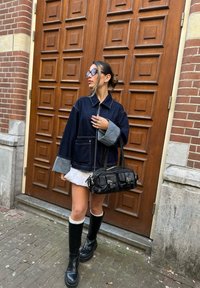 Donkerdenimjack met grijze manchetten, gedragen over een witte rok. Zwarte handtas met meerdere zakken. Zwarte kniehoge laarzen. Achtergrond met een bruine houten deur.