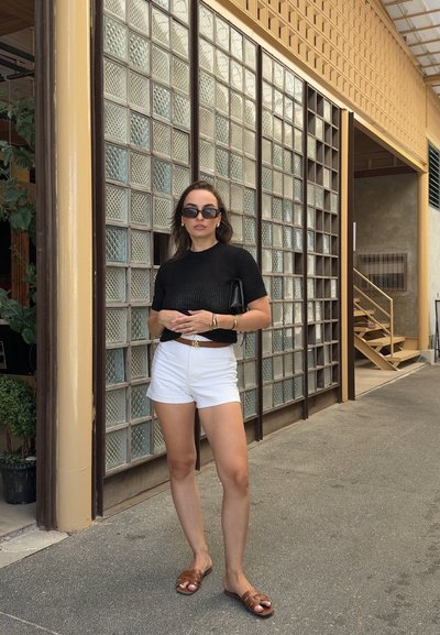 Top de punto negro de manga corta, pantalones cortos blancos de cintura alta, sandalias marrones y un bolso de mano negro. De pie frente a una pared de bloques de vidrio.