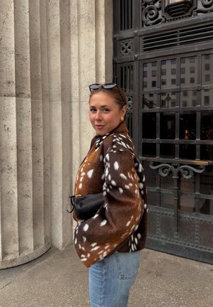 Mujer que lleva una chaqueta de piel con manchas marrones y blancas y vaqueros azules, de pie junto a altas columnas de piedra y una puerta de metal negro ornamentada.