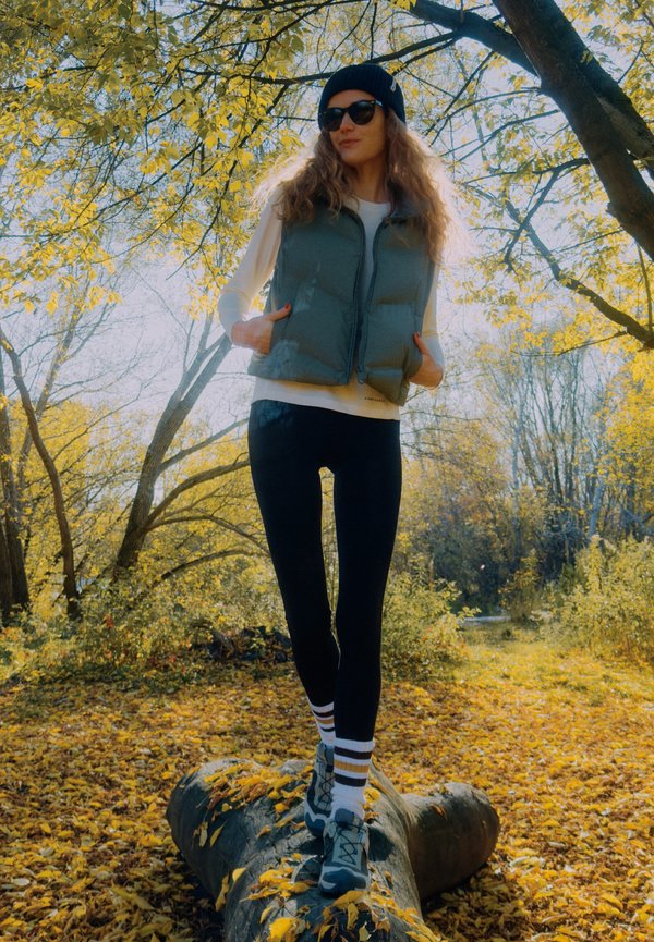 Veste matelassée grise sur une chemise blanche à manches longues, leggings noirs et chaussures grises avec chaussettes rayées, en plein air avec un feuillage d'automne.