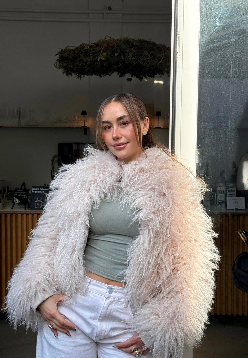 Jeune femme aux longs cheveux portant une veste moelleuse beige, un crop top vert clair et un pantalon blanc, debout dans l'intérieur d'un café moderne.