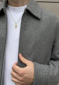 Grey wool-blend jacket with a zip front, collared design, and side pockets; paired with a white T-shirt and a gold wing pendant necklace.