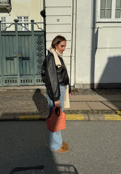 Chaqueta de cuero negra corta, bufanda crema, pantalones vaqueros acampanados azul claro y zapatos marrones. Sosteniendo un bolso de mano tejido de color coral.