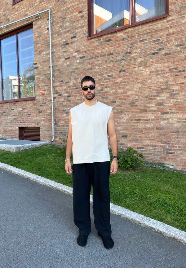 Cream sleeveless top, black loose pants, and black shoes. Standing against a brick wall with windows. Grass and sidewalk in the foreground.