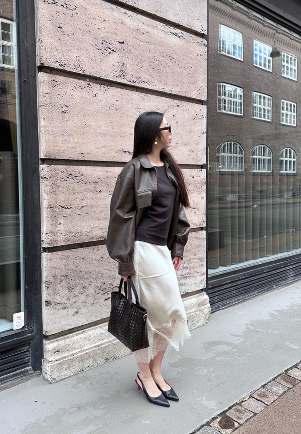 Mujer con chaqueta de cuero marrón, camiseta negra, falda blanca y tacones negros, sosteniendo un bolso negro, de pie en la acera cerca de una pared de un edificio.