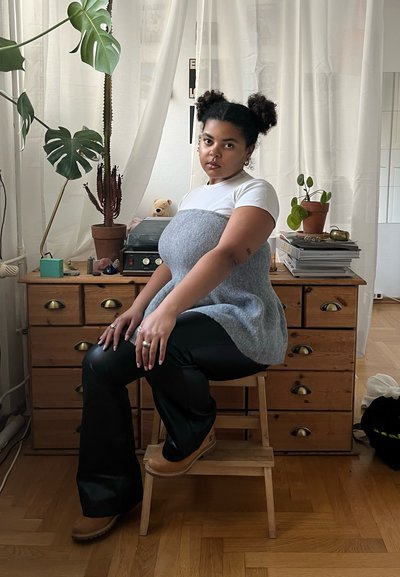 Túnica de punto gris sobre una camiseta blanca, pantalones de campana negros y zuecos color avena. Sentada en un taburete de madera, con plantas de interior y un cajón en el fondo.