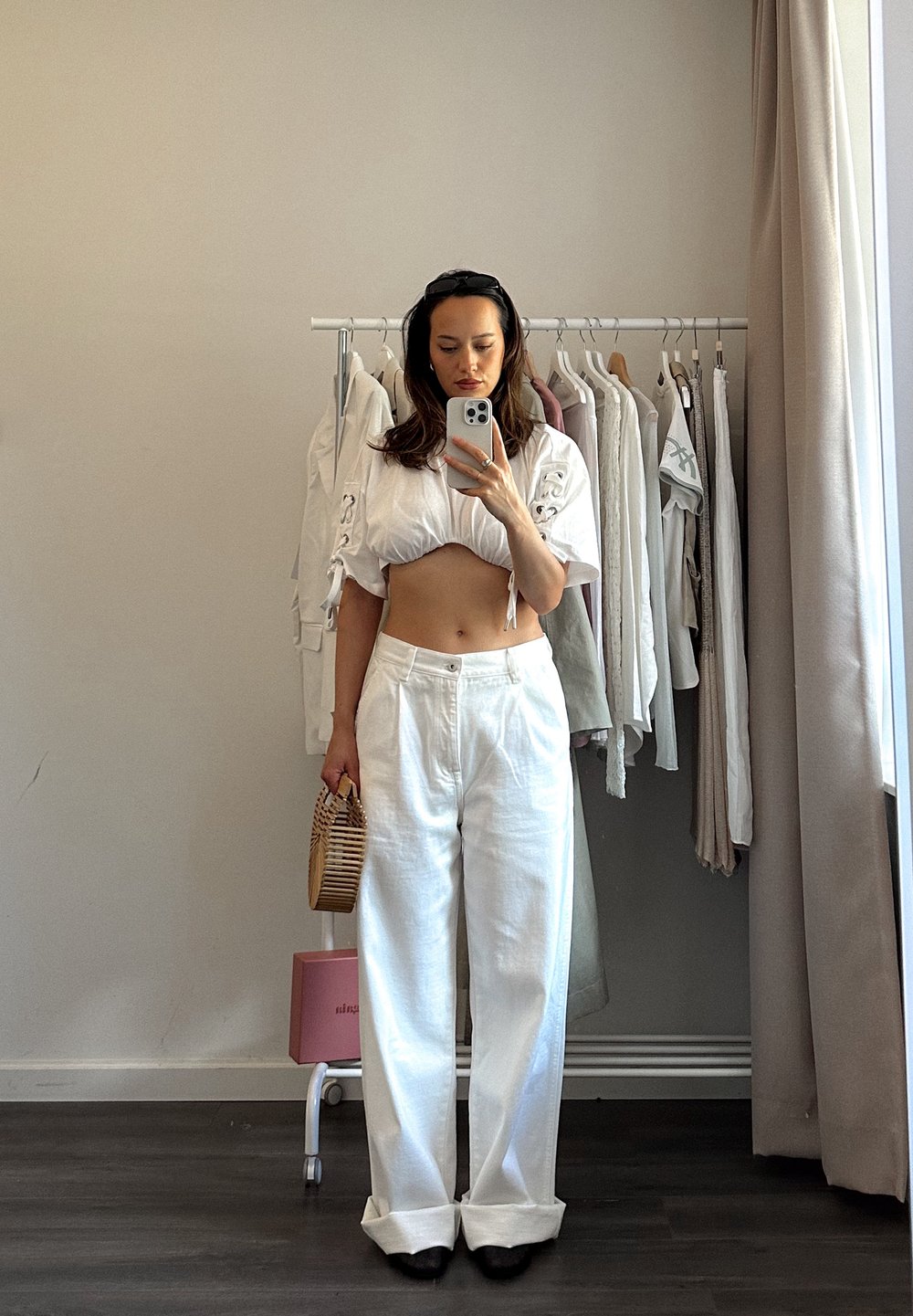 Λευκό κοντό τοπ με λεπτομέρεια δέσιμου μπροστά, ψηλόμεσα λευκά παντελόνια από λινό και μία ριγέ ψάθινη τσάντα; στέκεται μπροστά από μια κρεμάστρα ρούχων.