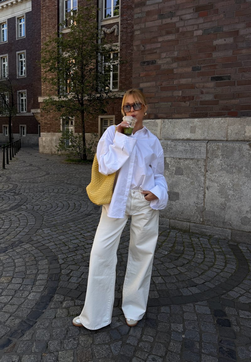 Femme portant une chemise blanche et un pantalon large, des lunettes de soleil, tenant un verre vert et un sac jaune, debout dans une rue pavée près d'un bâtiment en briques.
