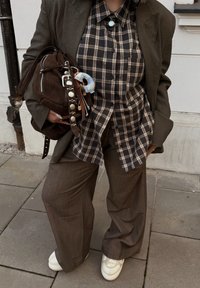 Blazer marron surdimensionné, chemise à carreaux noir et beige, sac marron avec différents éléments en métal, pantalon gris ample et baskets blanches.