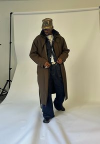 Mann in braunem Trenchcoat, Jeansjacke und -hose, weißem Hemd, Camouflage-Cap, Sonnenbrille und Schmuck, der in einem Studio mit weißem Hintergrund posiert.