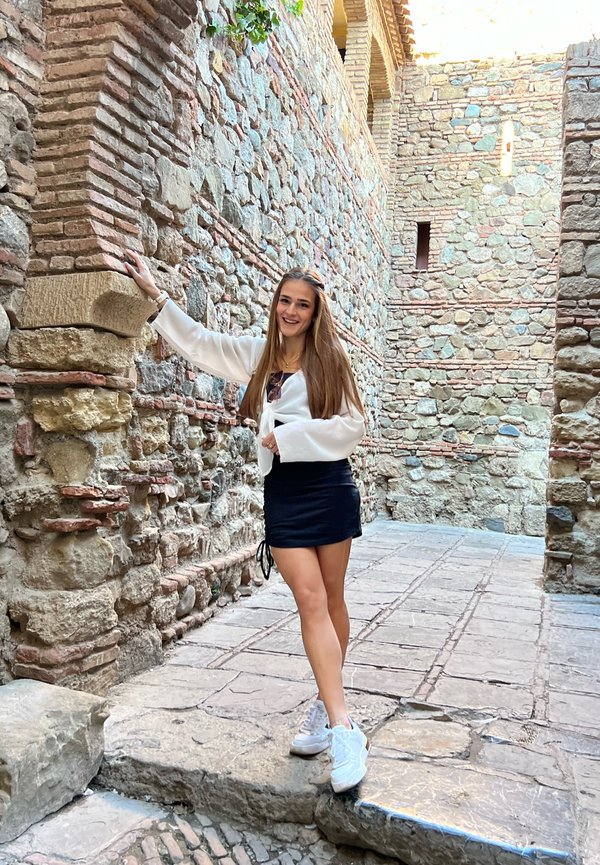 Jonge vrouw in een vloeiende witte blouse en zwarte rok poseert tegen een stenen muur, met een gestructureerd pad en groen in een historische omgeving.