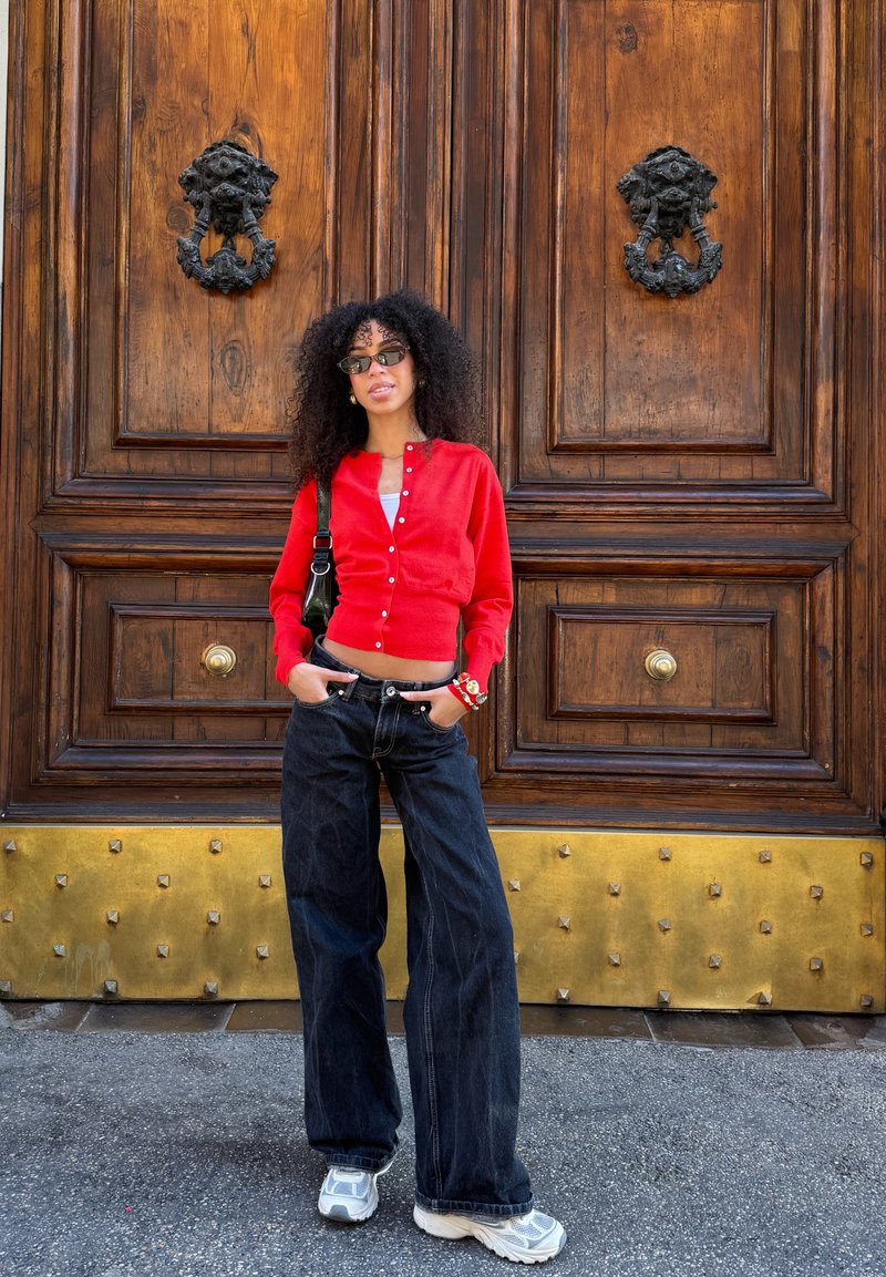 Femme aux cheveux bouclés portant des lunettes de soleil, un cardigan rouge, un jean large noir et des baskets blanches se tient devant de grandes portes en bois ornées.