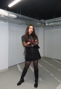 Crna kratka haljina s prozirnim slojem i šarenim grafičkim uzorkom, u kombinaciji s ribljom mrežom i crnim čizmama do koljena. Pozadina prikazuje valovite zidove.