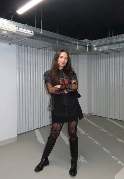 Vestido corto negro con superposición transparente y gráfico colorido, combinado con medias de red y botas negras hasta la rodilla. El fondo presenta paredes corrugadas.