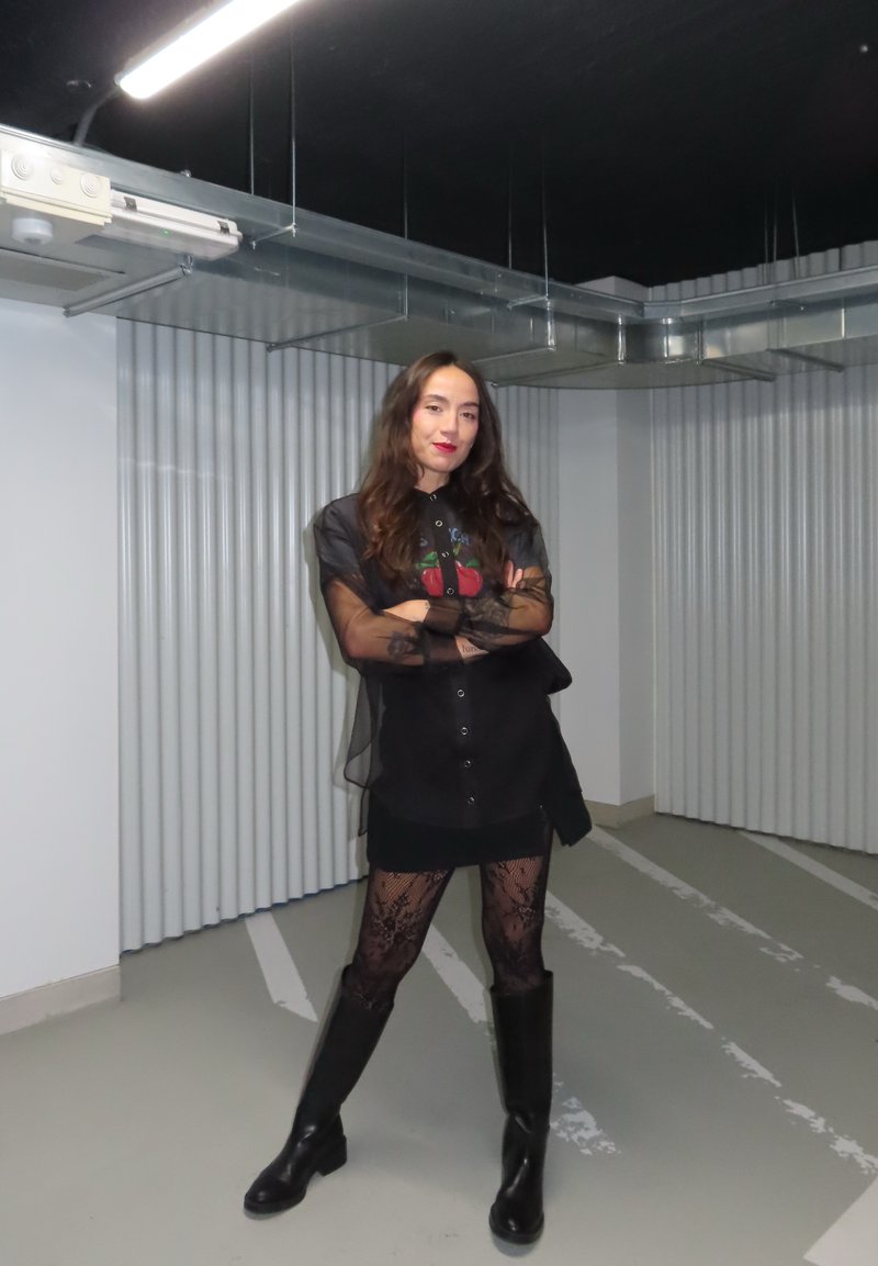 Must must outfit, millehüljed ja värviline graafika, koos kaltsujalatsite ja põlvekõrguste mustade saapadega. Taustal on lainelised seinad.