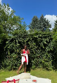 Un vestido rojo con una tela texturizada, de pie sobre hierba verde cerca de una manta blanca. Toalla blanca en mano, rodeada de vegetación y un bol de fresas.