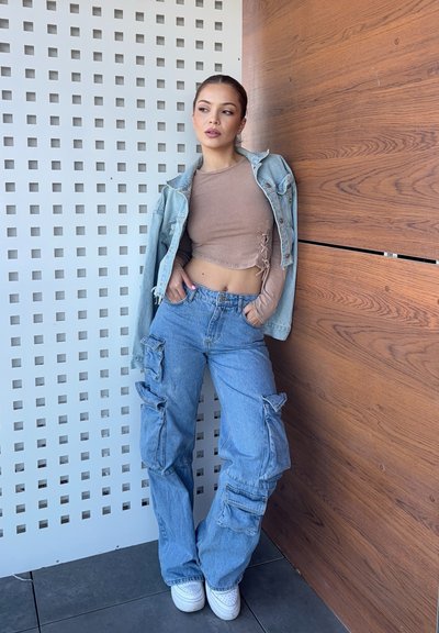 Pantalones cargo de mezclilla azul claro con múltiples bolsillos, combinados con un top corto marrón y una chaqueta de mezclilla clara reposando sobre el hombro.