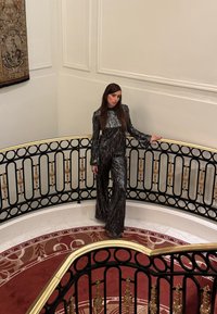 Femme portant une combinaison rayée noire et argent, appuyée sur une rampe ornée noire et dorée dans une pièce blanche avec un tapis rouge à motifs.