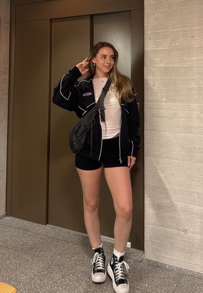 Joven mujer de pie en el interior junto a un ascensor, con pantalones cortos negros, chaqueta negra, top claro, zapatillas negras altas y bolso cruzado.