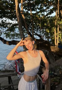 Muskuļots cilvēks pelēkā bezroku krekliņā un baltos šortos, stāv pie koka pie ūdens, turdams bundžu un taisot rotaļīgu žestu.