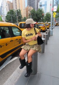 Gul grafisk t-skjorte med "AVAYAV" tekst, camo miniskjørt, svarte gummistøvler og en beige baseballlue. Urban setting med gule drosjer.