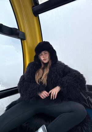 Joven mujer con abrigo peludo negro y sombrero, sentada dentro de una cabina de góndola amarilla con ventanas empañadas, las manos descansando sobre su regazo.