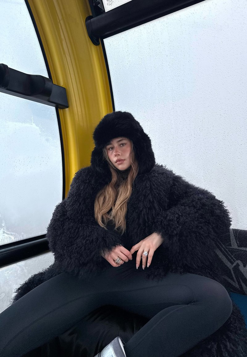 Ung kvinne i en svart pelsaktig jakke og hatt sitter inne i en gul gondolhytte med tåkete vinduer, hendene hvilende i fanget.