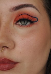 Primo piano di un volto con ombretto arancione e eyeliner blu a forma di ala, un anello nasale dorato e un colore labbra opaco rossastro. La pelle appare liscia.