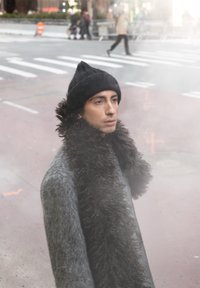 Jeune personne portant un bonnet en tricot noir et une écharpe sombre et duveteuse, debout près d'un passage pour piétons avec des piétons flous en arrière-plan.