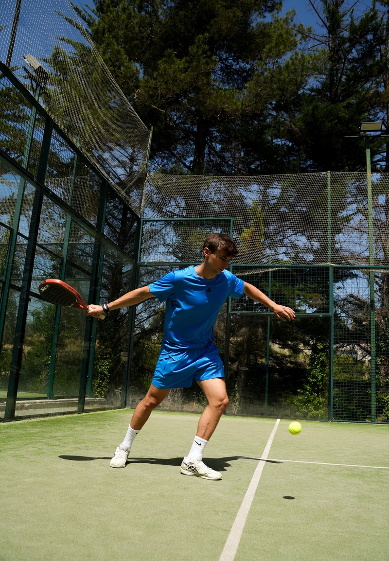 Mladý muž v modrom športovom oblečení hrá padel, pripravuje sa na úder loptičky na vonkajšom kurte obklopenom zeleným plotom a stromami.