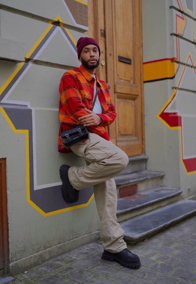 Camisa de franela a cuadros rojos y naranjas, pantalones cargo beige, zapatos de plataforma negros y un bolso negro pequeño. El fondo presenta un mural colorido y una puerta de madera.