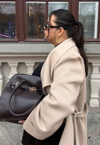 Bézs kabát, fekete felső, nagy barna bőrtáska arany csattal és fogantyúval. Hosszú, egyenes haj hátrafogva, fülbevaló karikákkal.