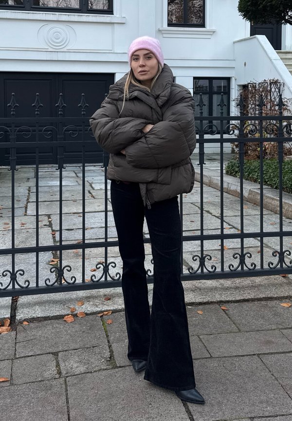 Veste matelassée marron foncé, pantalon noir évasé et un bonnet rose pâle. Debout devant une grille noire et un bâtiment blanc moderne.