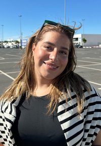 Sciarpa a righe bianche e nere sopra una maglietta nera, con capelli castani alla spalla, sorridente contro uno sfondo di cielo blu limpido e un parcheggio.